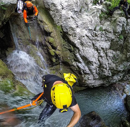 Canyoneering member with backpack rappeling down the waterfall in the canyon. Other member nearby is making a video.
